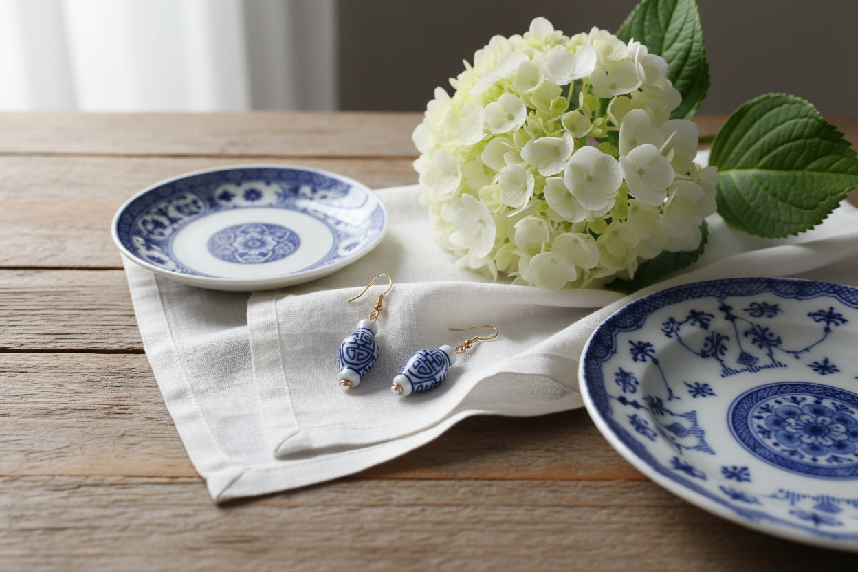 A pair of dangling earrings with small white and blue patterned ceramic vase charms and gold fishhook wires, displayed against a light gray background.
