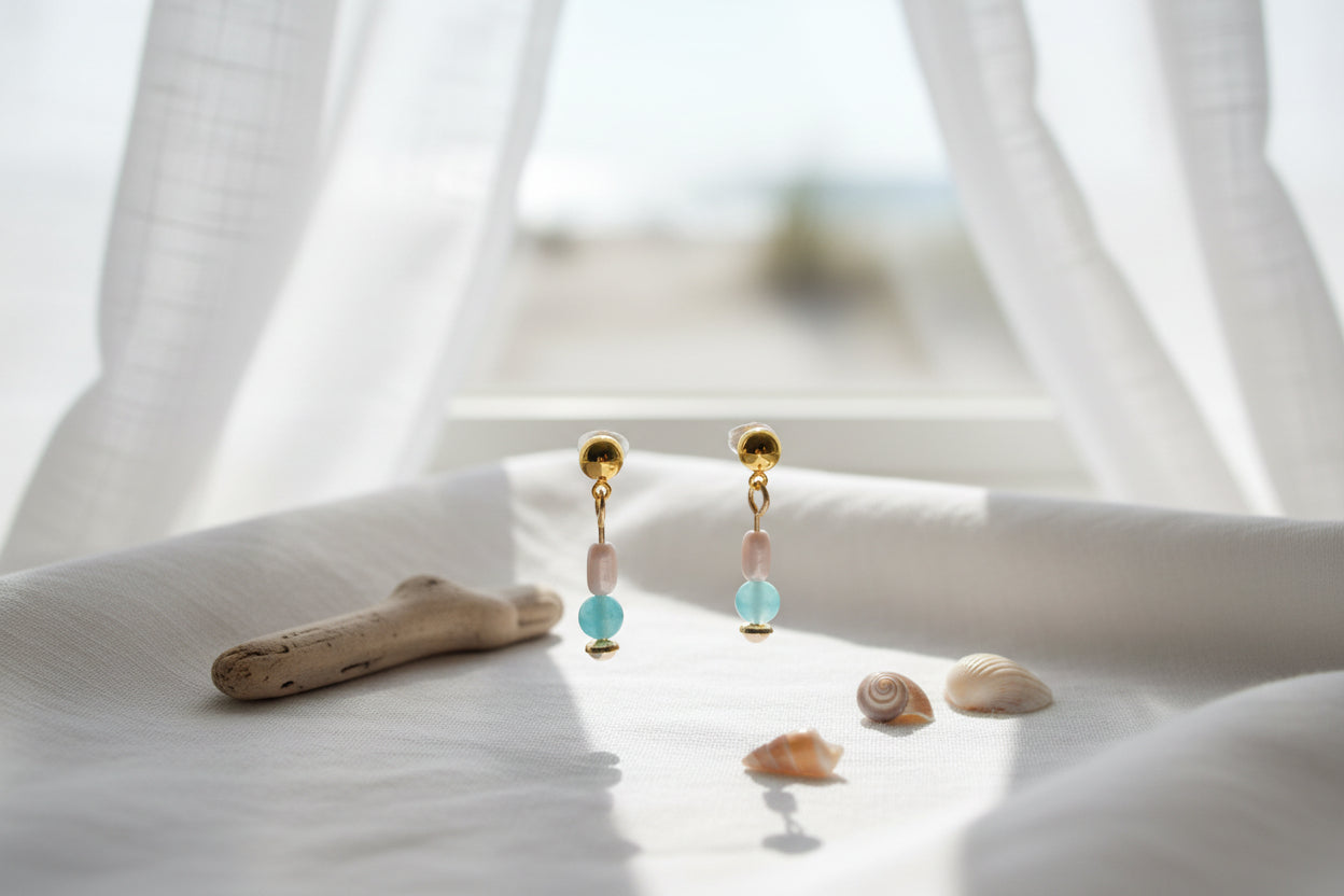 A pair of gold dangle earrings with a white bead, a light blue bead, and a small gold disc at the bottom, displayed on a clear acrylic stand against a light gray background.
