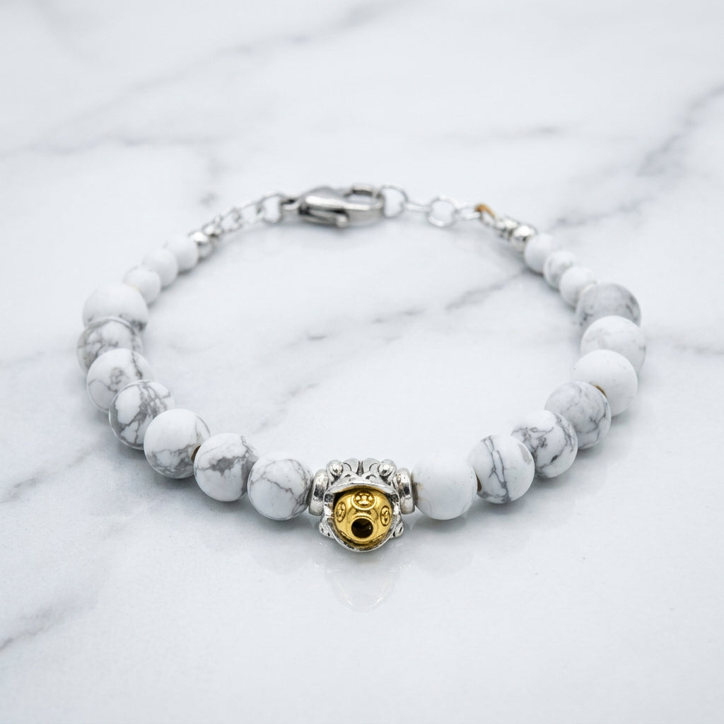 A beaded bracelet featuring light gray and white marbled beads with a central gold and silver charm, displayed on a neutral gray background.