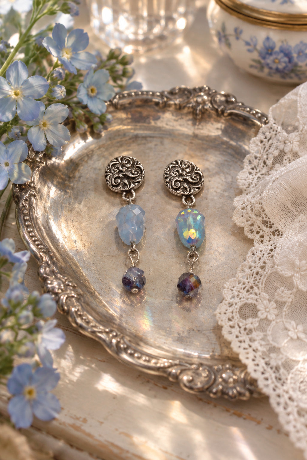 A pair of dangling earrings on a white display card. The earrings have ornate silver-toned filigree studs, a light blue faceted bead, an iridescent white bead, and a small dark brown bead at the bottom.