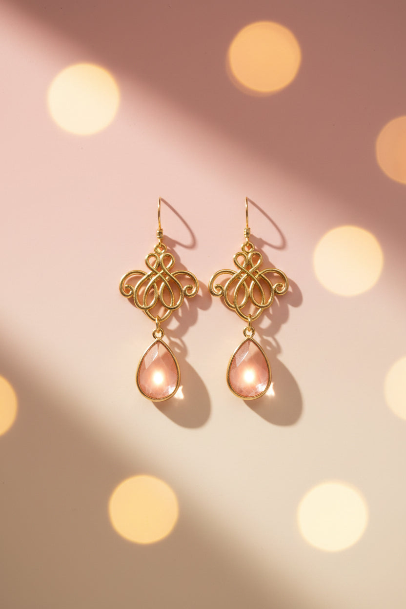 A pair of gold-tone drop earrings with ornate filigree tops and teardrop-shaped pink gemstones, displayed on a white card labeled 'River Inspired Handcrafted Jewelry' against a plain light gray background.