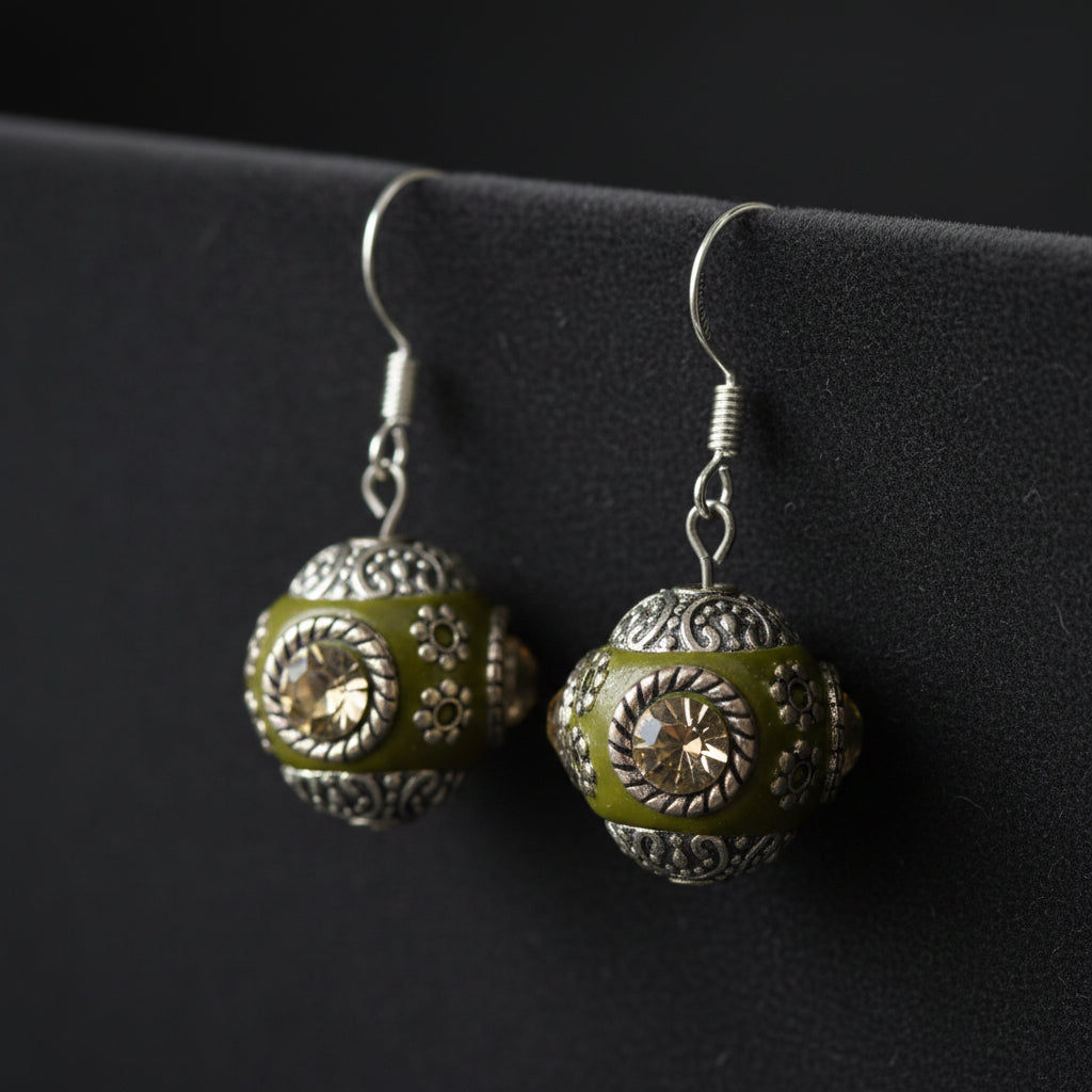 A pair of handmade dangle earrings with olive green enamel and silver filigree, hanging from silver fishhook wires on a clear acrylic display stand against a neutral gray background.