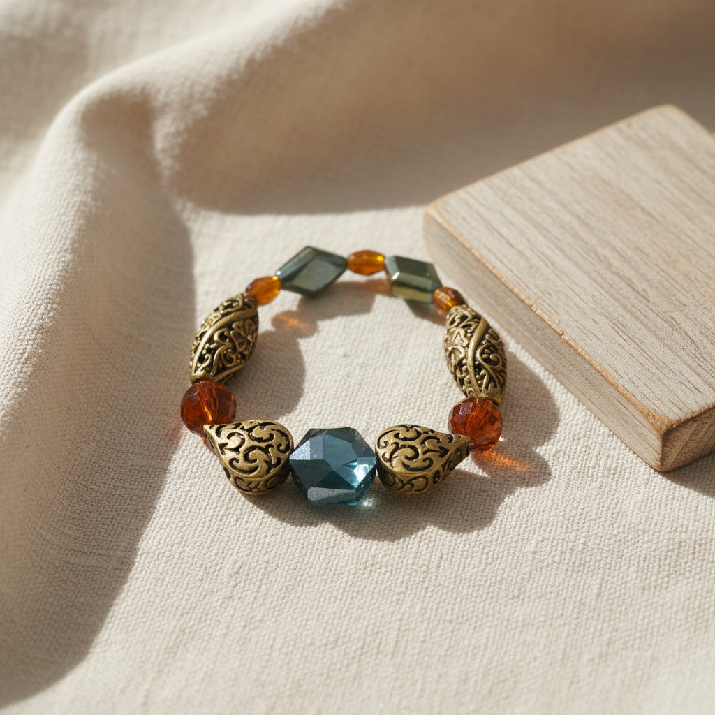 A beaded stretch bracelet featuring ornate gold filigree beads, translucent amber-orange beads, and a central faceted dark blue-green bead, displayed on a plain light gray background.
