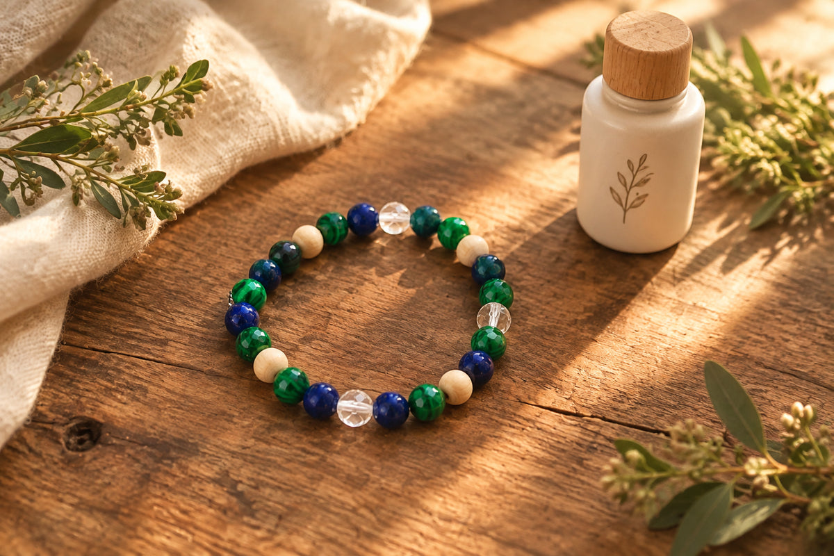 A beaded stretch bracelet featuring dark blue lapis lazuli, emerald green malachite, light beige wooden beads, and a clear faceted crystal bead, displayed on a neutral gray background.