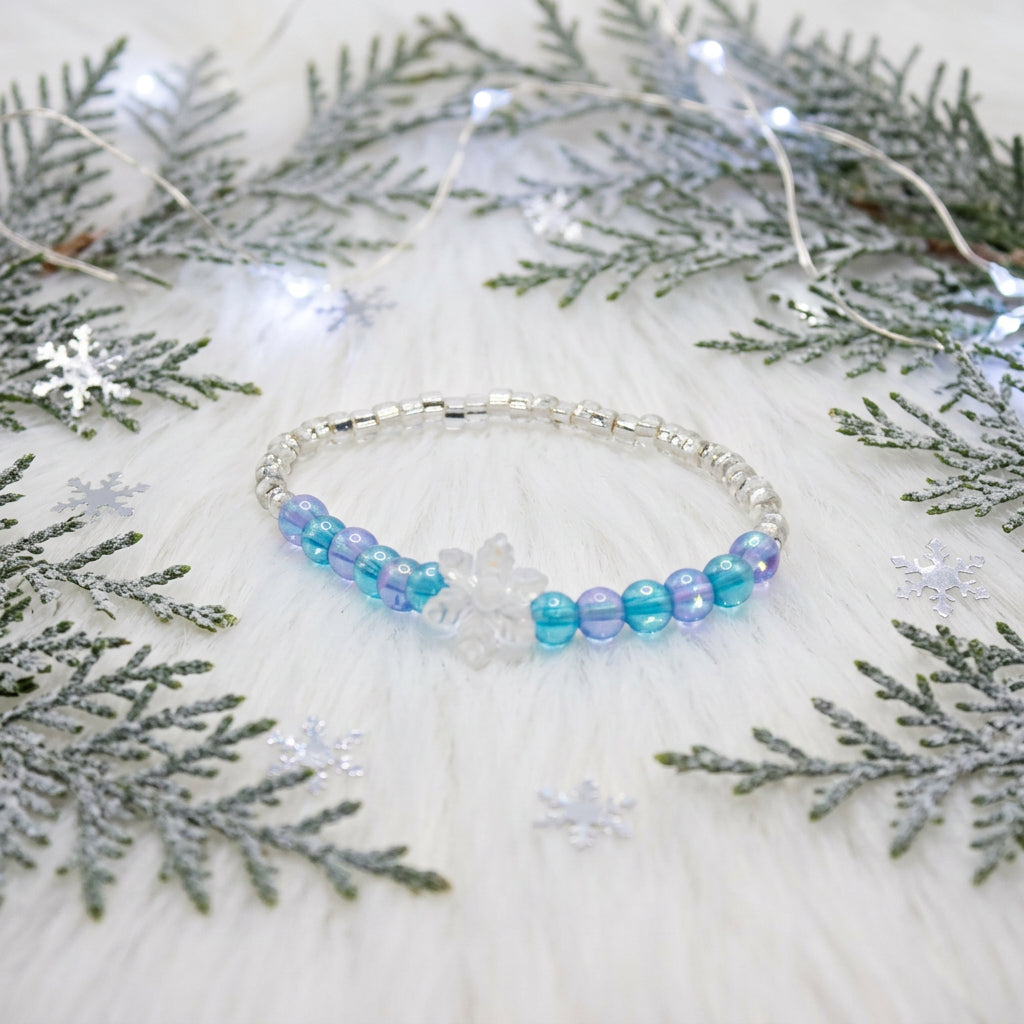 A beaded stretch bracelet featuring silver-toned metal beads, translucent blue beads, light blue beads, and a small white flower charm, displayed on a plain light gray background.