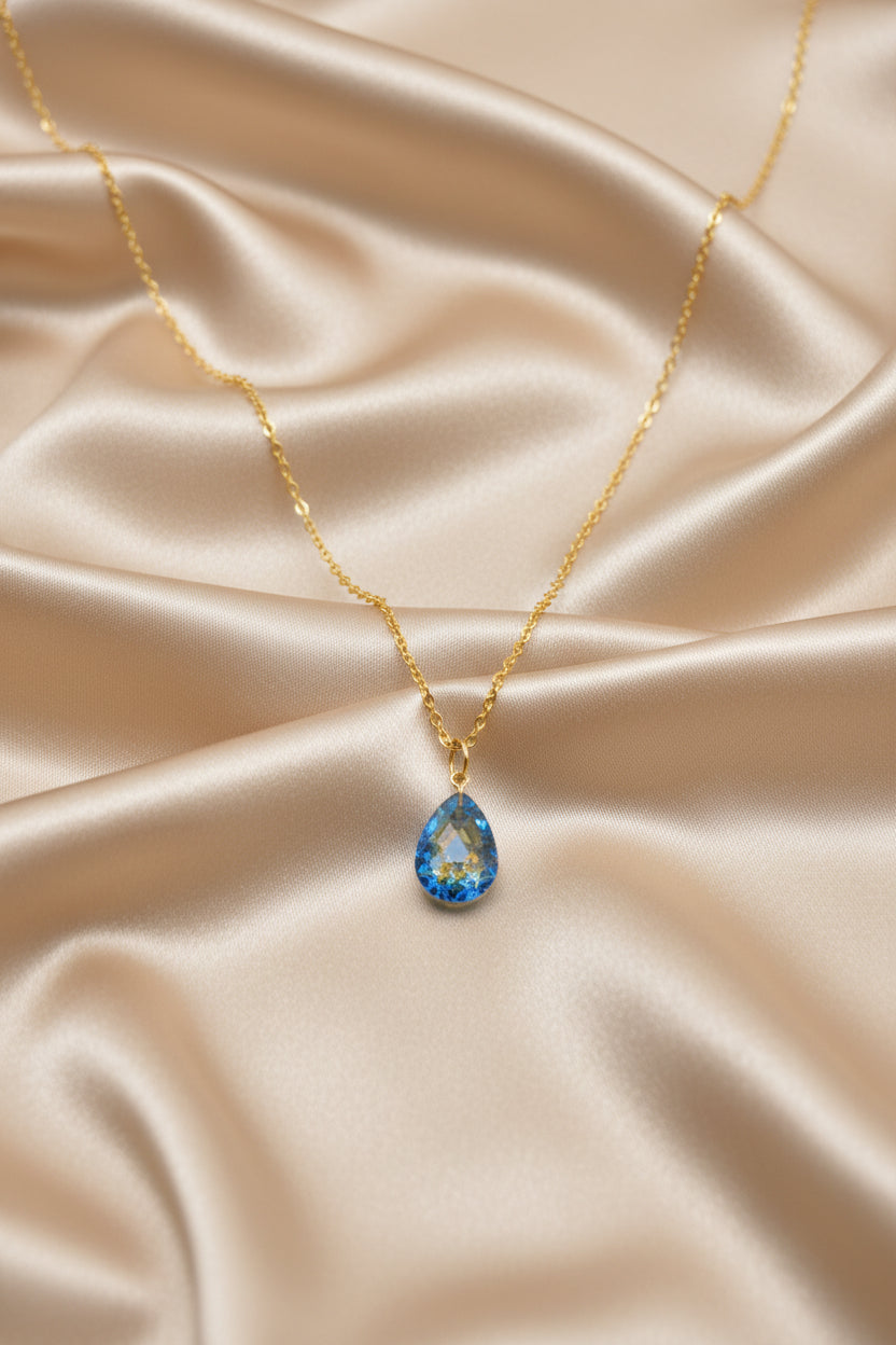 A gold chain necklace with a small teardrop-shaped pendant featuring a blue and gold pattern, displayed on a black velvet jewelry bust against a white background.