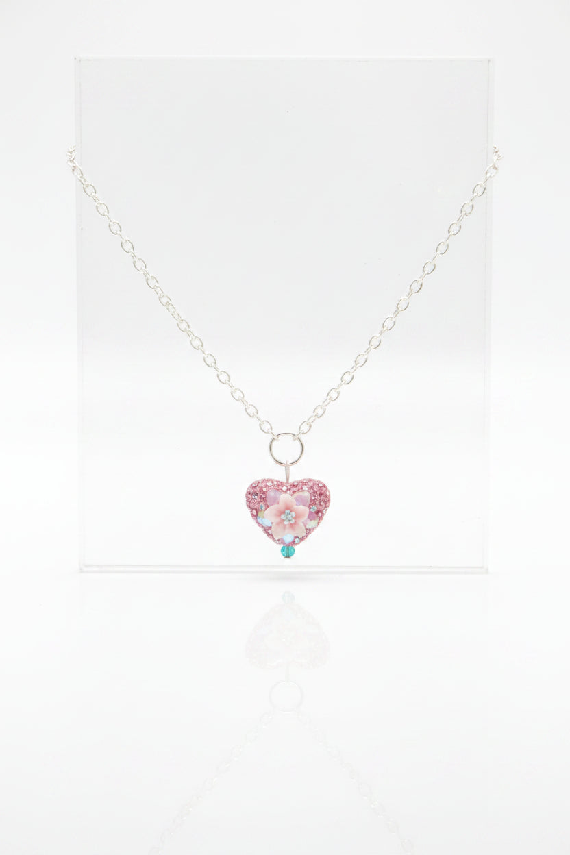 A silver chain necklace with a small heart-shaped pink rhinestone pendant, displayed on a black velvet jewelry bust against a white background.