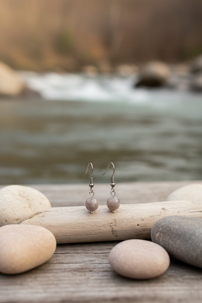 A pair of handmade dangle earrings with brown and gray beads on silver hooks, displayed on a white card labeled 'River Inspired Handcrafted Jewelry' against a neutral gray background.