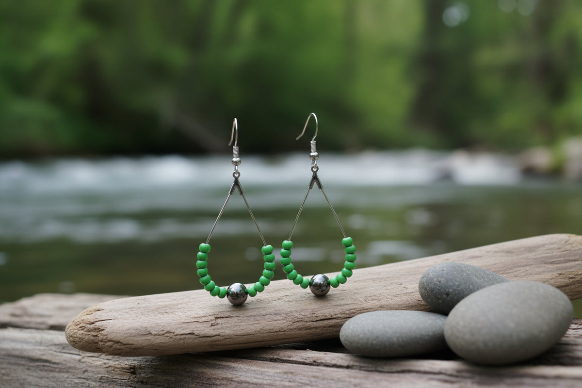 A pair of handmade dangle earrings with green faceted beads forming a semicircle, a central dark brown bead, and silver-toned fishhook wires, displayed on a clear acrylic stand against a neutral gray background.