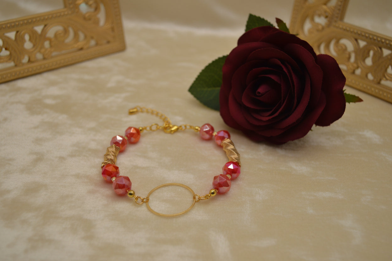 A gold-tone beaded bracelet with pink and red marbled beads and a central open circle charm, displayed on a light gray background.