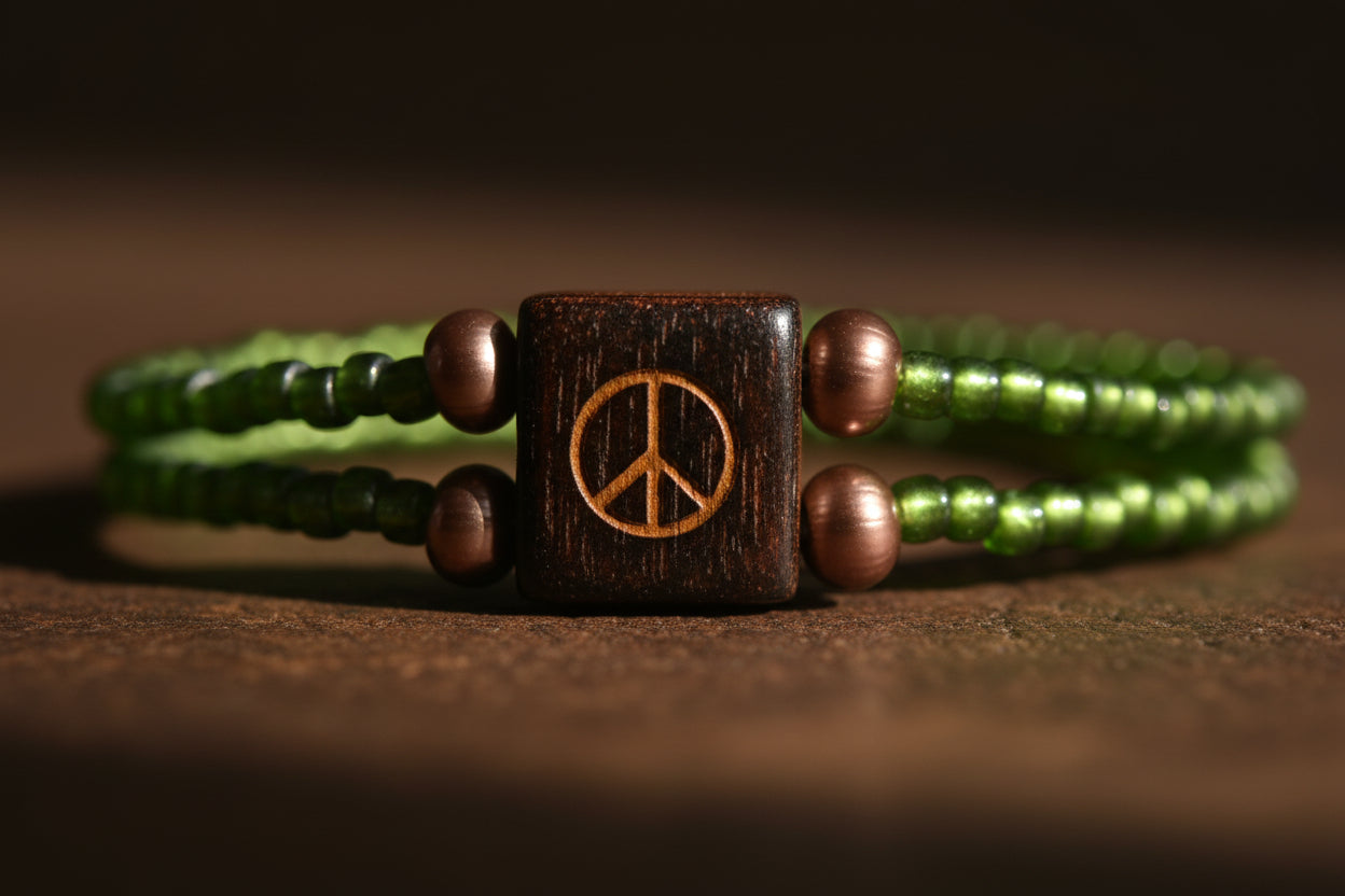 A beaded wrap bracelet with green beads, small metallic bronze beads, and a central dark brown square charm featuring a gold peace sign, displayed on a plain light gray background.