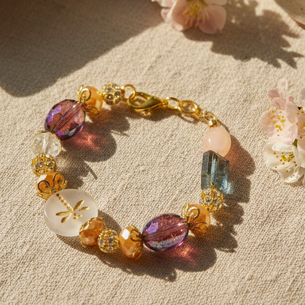 A beaded bracelet with a gold-tone clasp, featuring iridescent purple oval beads, peach-colored round beads, gold rose-shaped beads, and dark green rectangular beads on a light gray background.