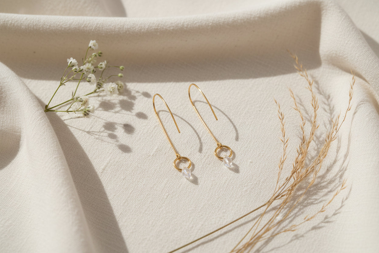A pair of minimalist gold dangle earrings with thin vertical bars, circular wire hoops, and clear teardrop crystal pendants, displayed on a clear acrylic stand against a neutral gray background.