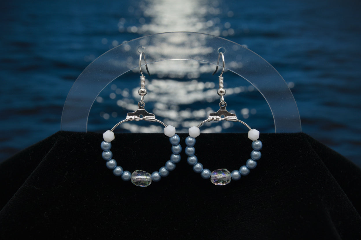A pair of handmade hoop earrings with blue and white beads, silver-tone metal hoops, and silver fishhook ear wires, displayed on a clear stand against a light gray background.