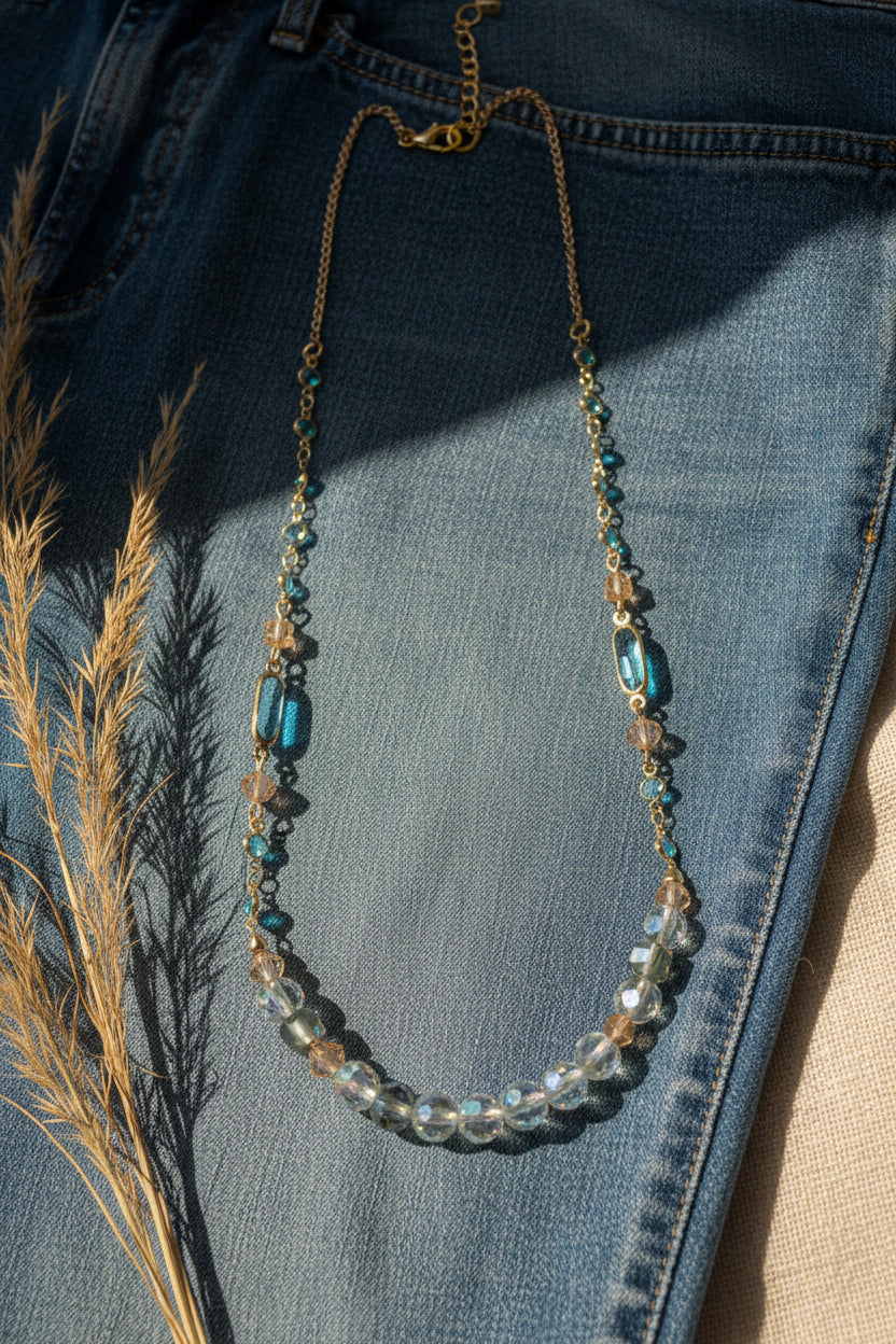 A delicate gold chain necklace featuring a graduated bead strand with clear faceted beads, light blue oval beads, and small iridescent beads, displayed on a neutral gray background.