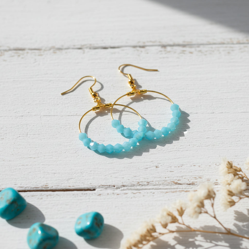 A pair of gold hoop earrings with small faceted light blue beads along the bottom rim, displayed against a neutral light gray background.