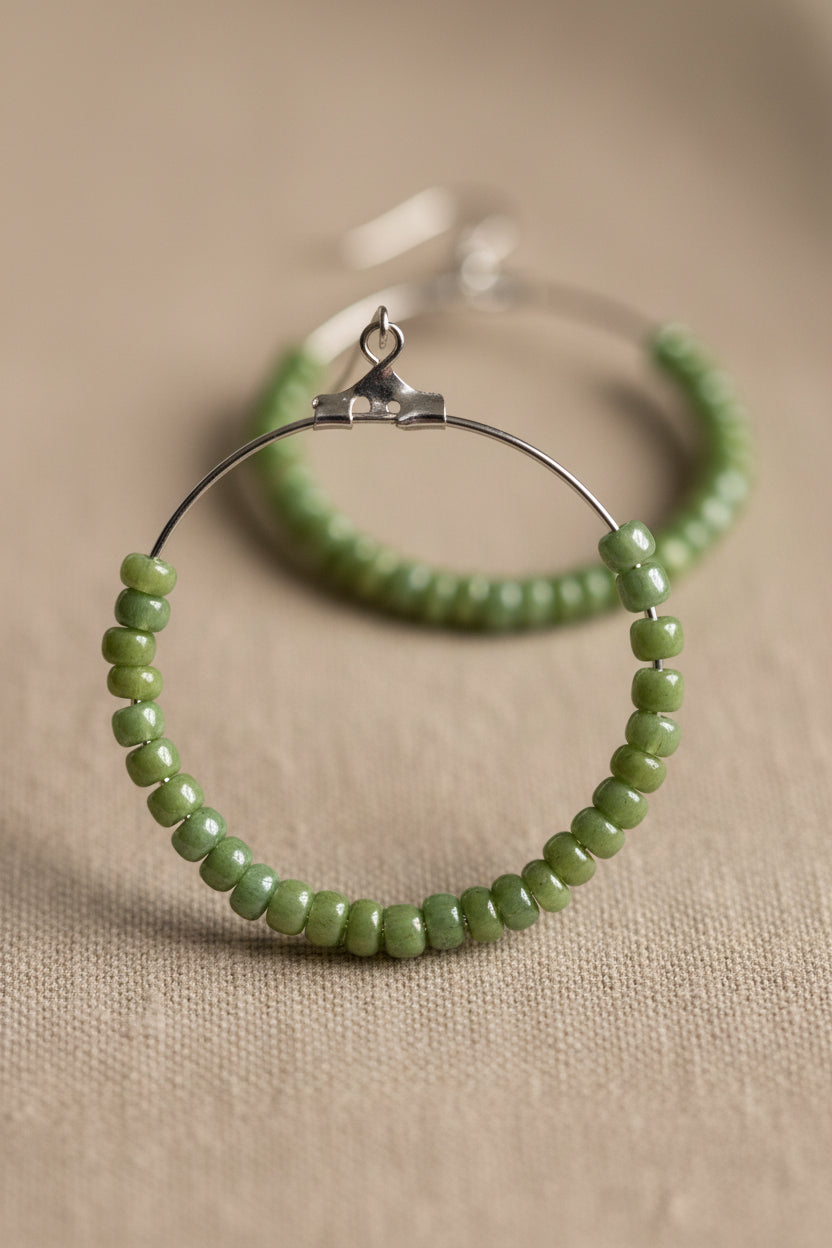 Green beaded hoop earrings on white display cards from River Inspired Handcrafted Jewelry, placed on a light surface with a clear plastic bag behind them.