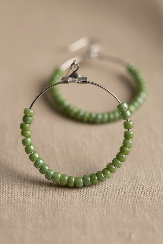 Green beaded hoop earrings on white display cards from River Inspired Handcrafted Jewelry, placed on a light surface with a clear plastic bag behind them.