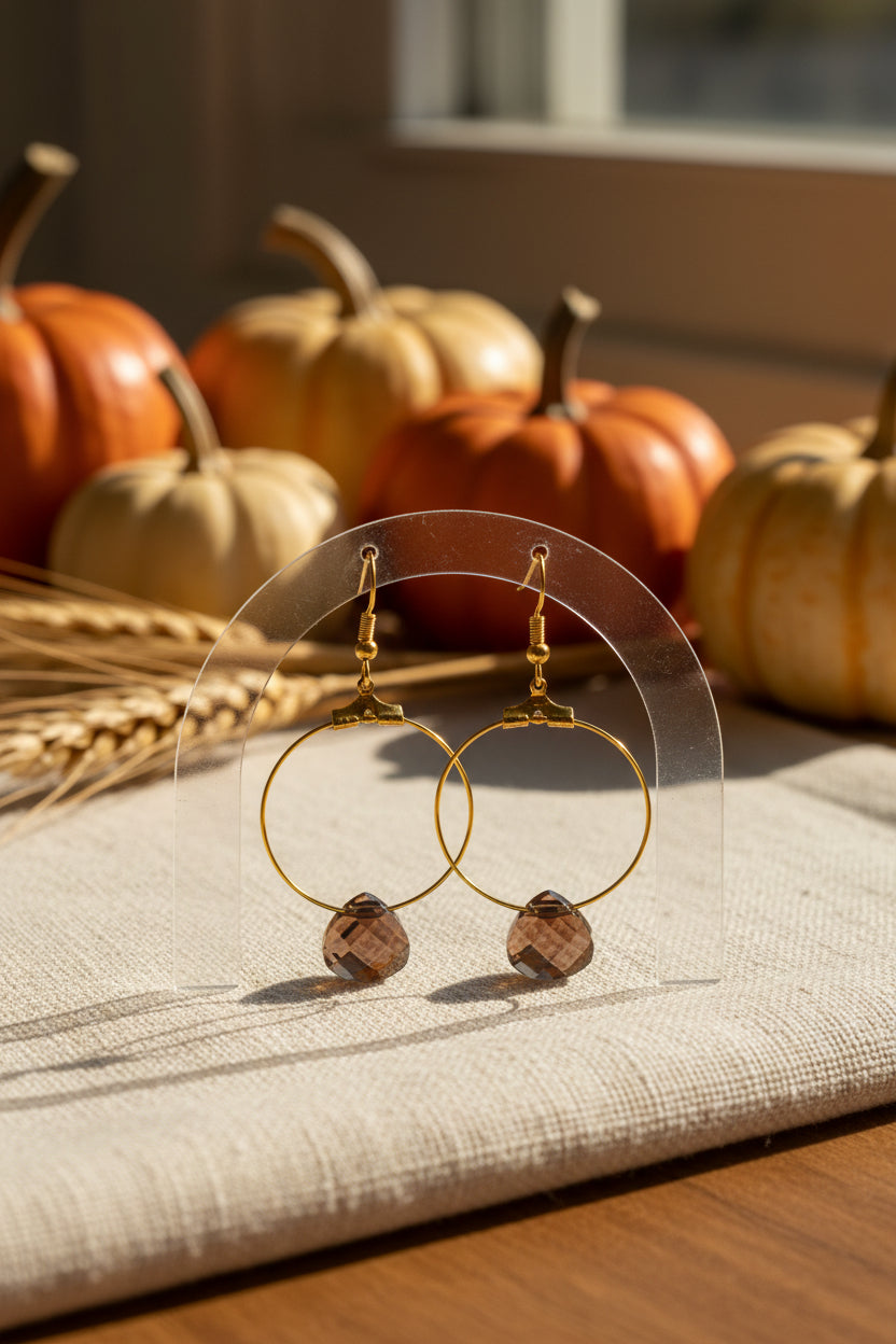 A pair of gold hoop earrings with faceted brown gemstone pendants, displayed against a neutral light gray background.