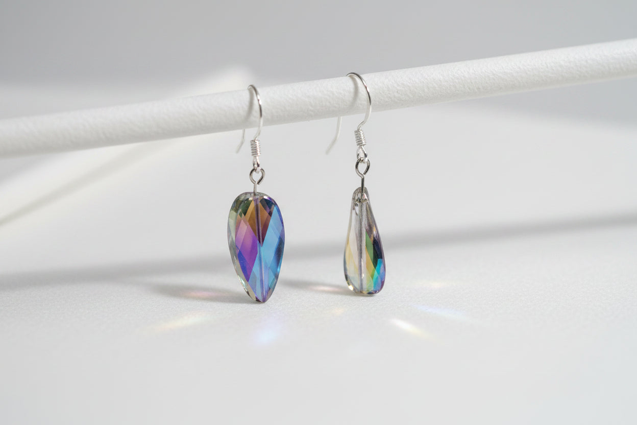 A pair of teardrop-shaped iridescent glass beads on silver fishhook ear wires, displayed on a clear plastic stand against a light gray background.