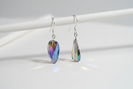 A pair of teardrop-shaped iridescent glass beads on silver fishhook ear wires, displayed on a clear plastic stand against a light gray background.