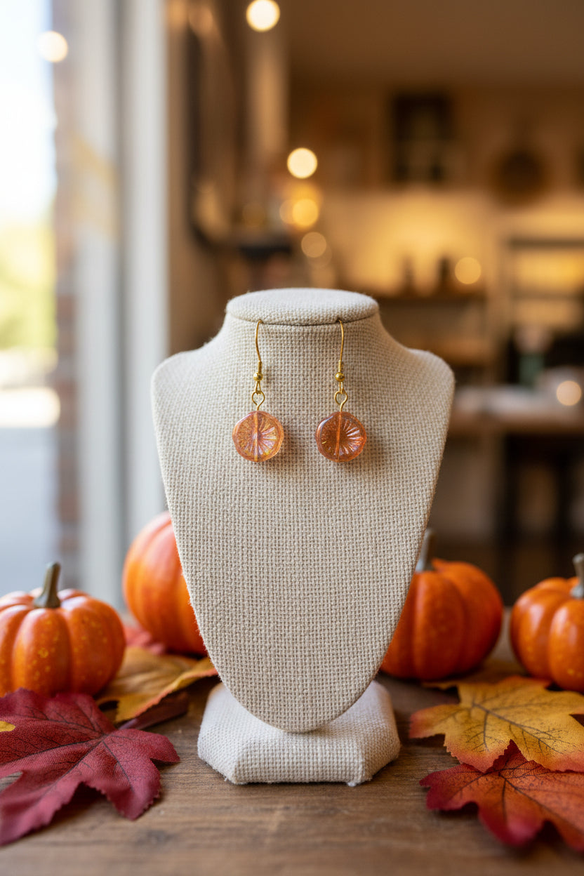 A pair of gold-tone leverback earrings with round, textured amber-orange glass pendants, displayed on a clear acrylic stand against a neutral gray background.
