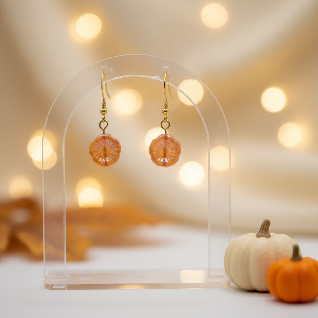 A pair of gold-tone dangle earrings with round, textured orange and peach resin pendants, displayed on a clear acrylic stand against a neutral gray background.