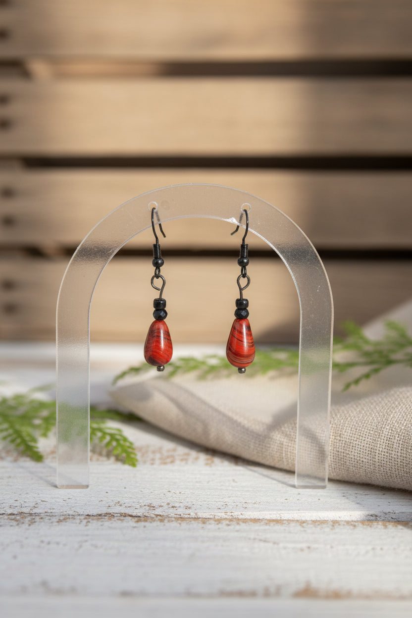 A pair of handmade dangle earrings with teardrop-shaped red marbled beads and black metal hooks, displayed on a clear plastic stand against a neutral gray background.
