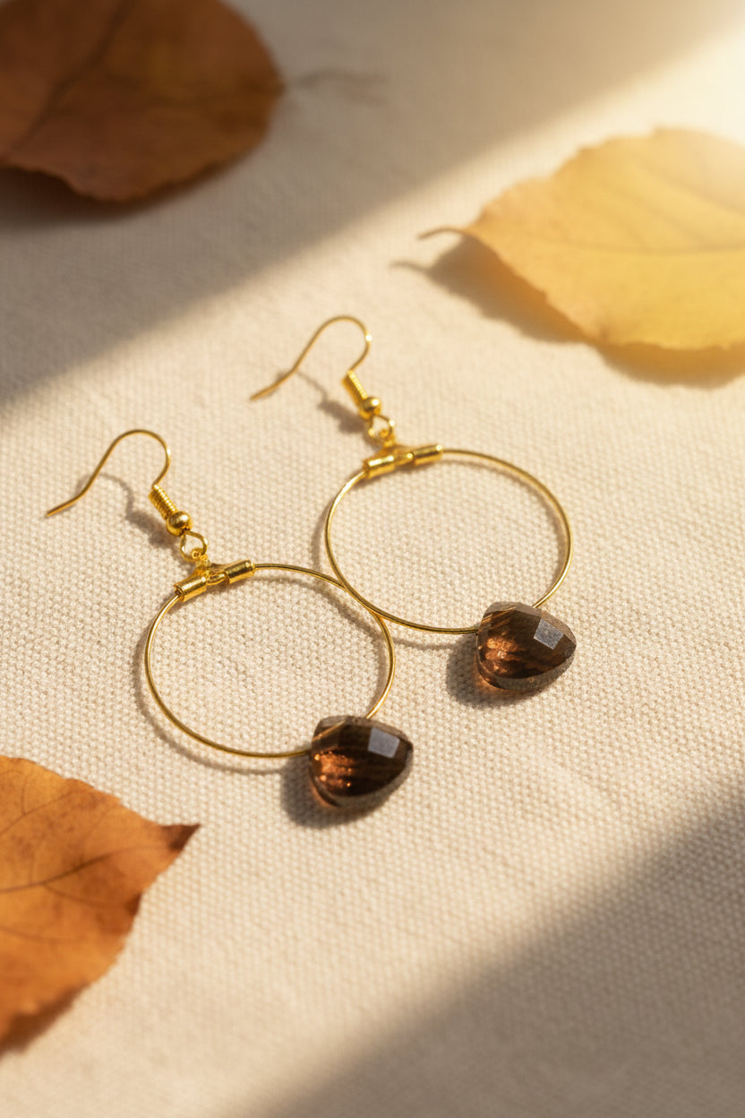 A pair of gold hoop earrings with faceted brown heart-shaped drop charms, displayed against a light gray background.