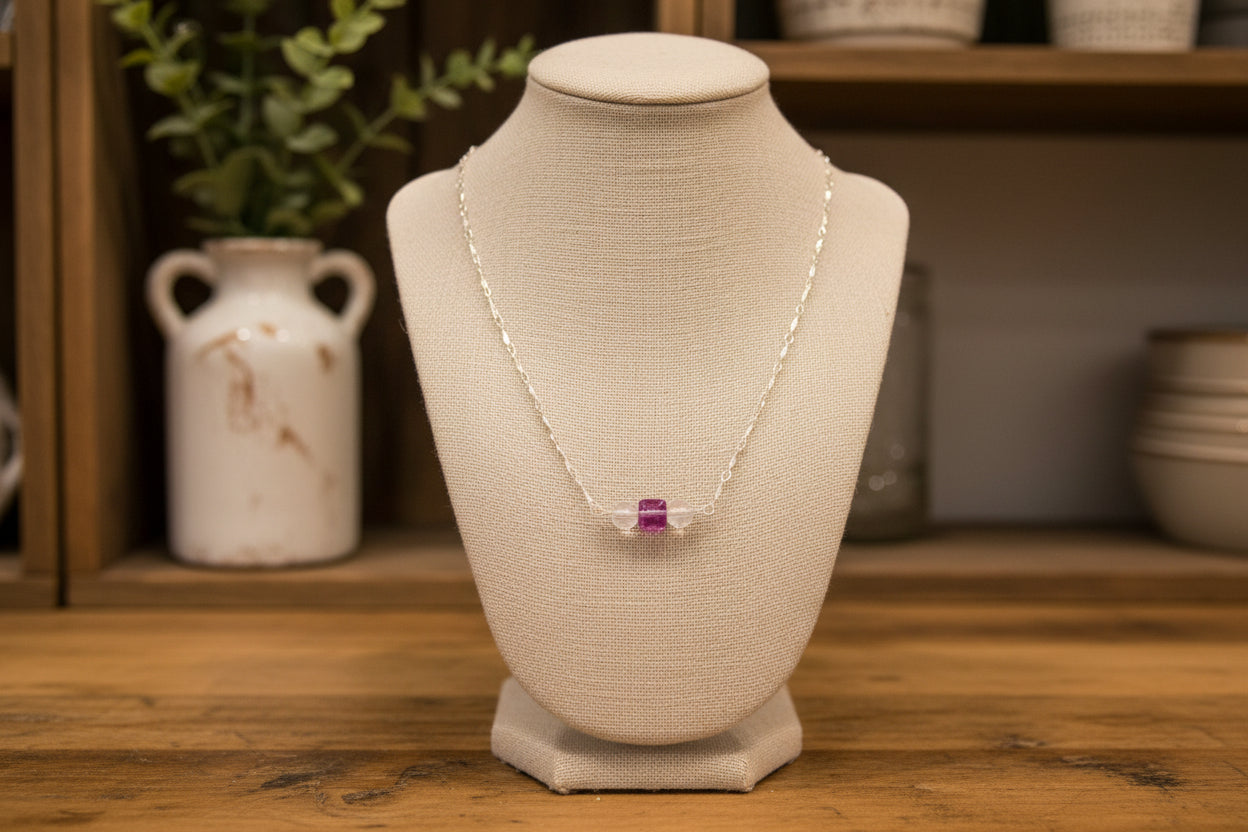 A silver chain necklace with three small beads at the center, including two clear round beads and one pinkish-purple bead, displayed on a black velvet jewelry bust.