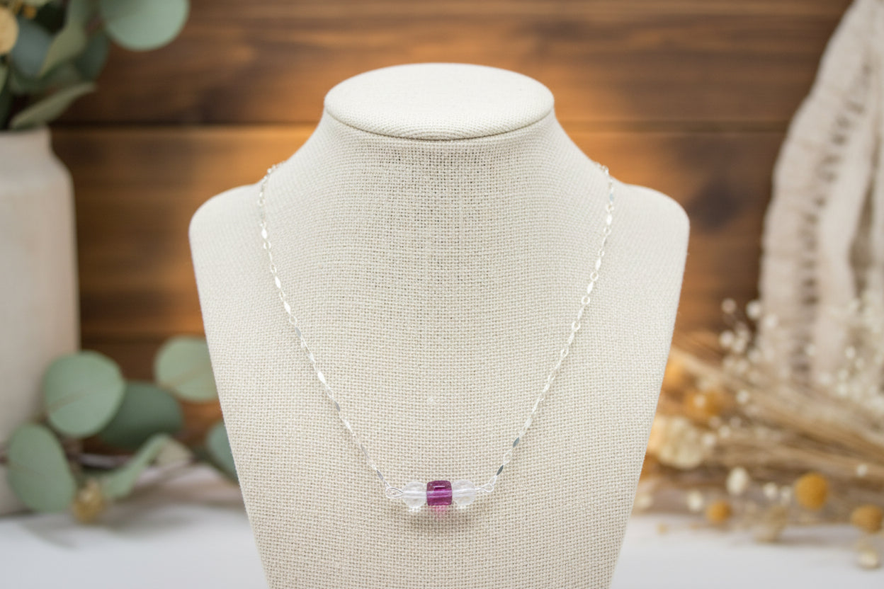 A silver chain necklace with three clear faceted beads and a small pink center bead, displayed on a black velvet jewelry bust against a white background.
