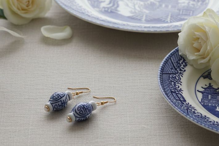 A pair of dangling earrings with small blue-and-white patterned ceramic vase charms and gold fishhook wires, displayed against a light gray background.