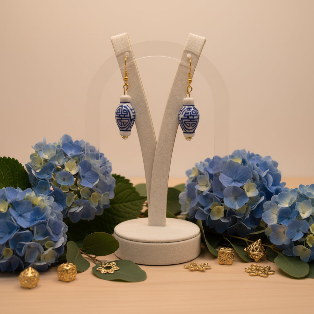 A pair of dangling earrings with small white and blue patterned ceramic vase charms and gold fishhook wires, displayed against a light gray background.