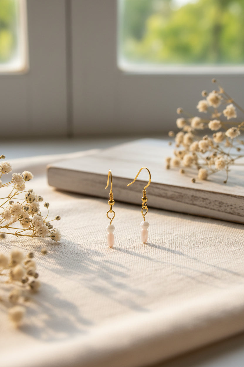 A pair of gold-tone dangle earrings with small white beads and pale pink stone drops, displayed against a light gray background.
