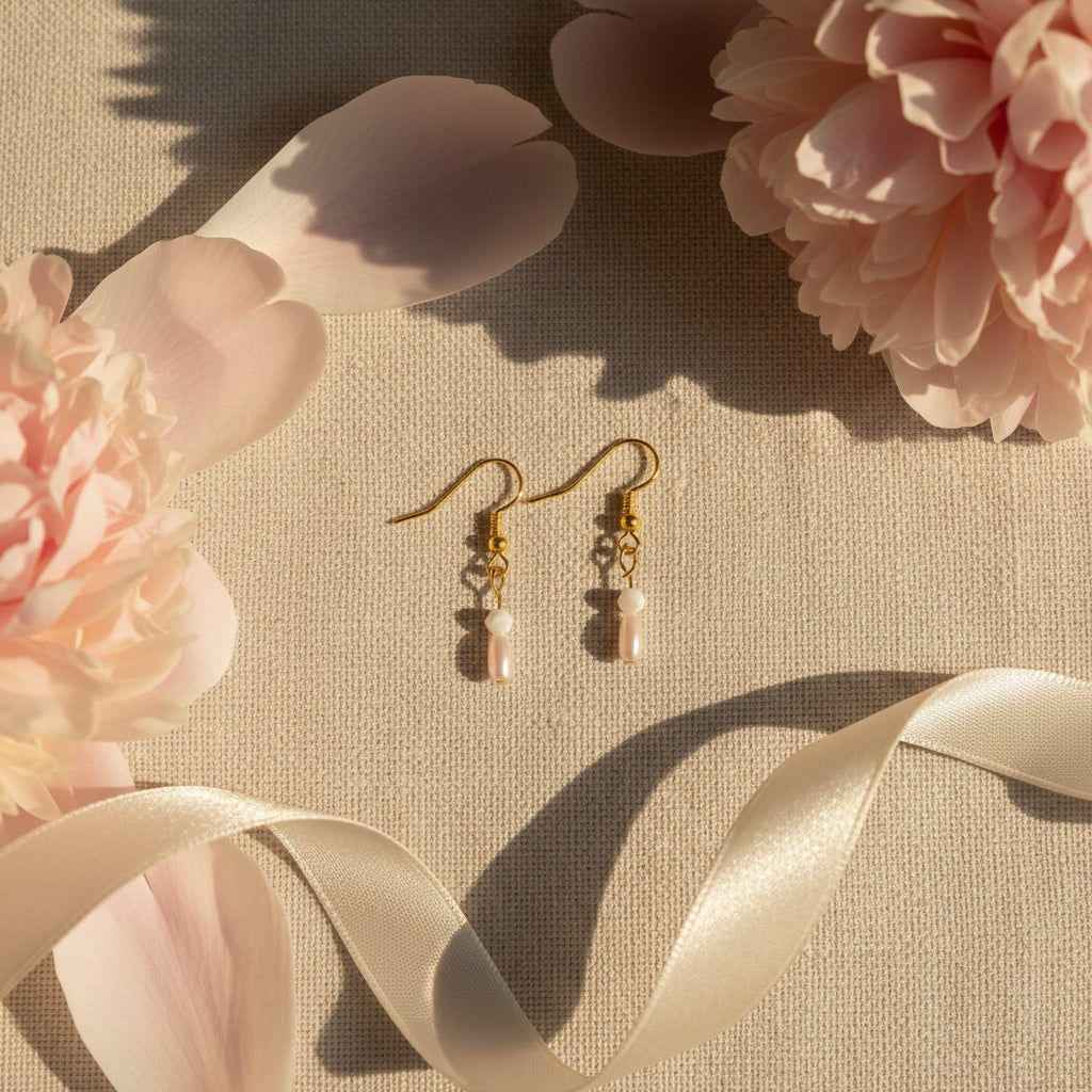 A pair of gold dangle earrings with small white beads and pale pink teardrop charms, displayed on a clear stand against a light gray background.