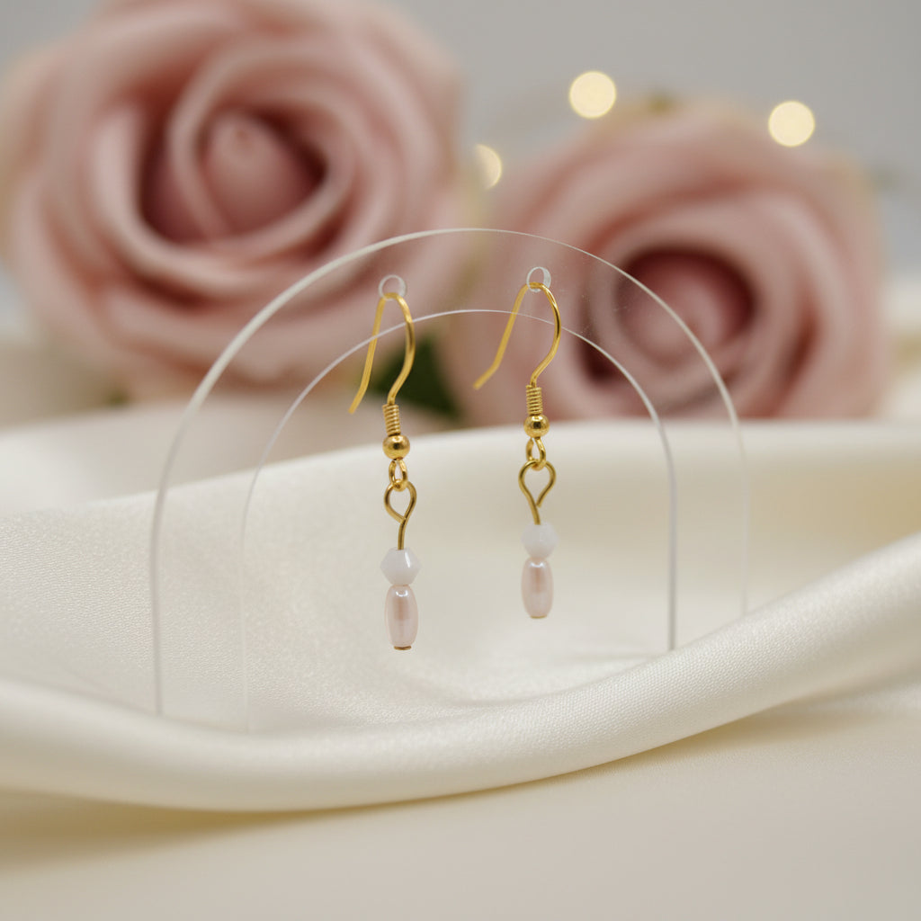 A pair of gold-toned dangle earrings with small white beads and pale pink teardrop charms, displayed against a plain light gray background.