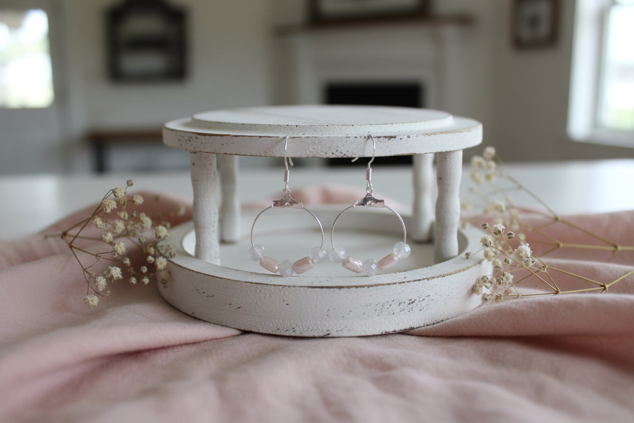 A pair of silver-tone hoop earrings with small pale pink and white beads along the bottom, displayed on a clear stand against a light gray background.