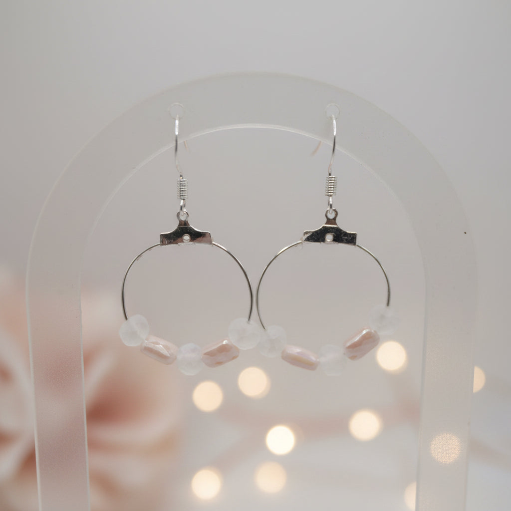 A pair of silver hoop earrings with small pale pink and white beads, hanging on a clear display stand against a light gray background.