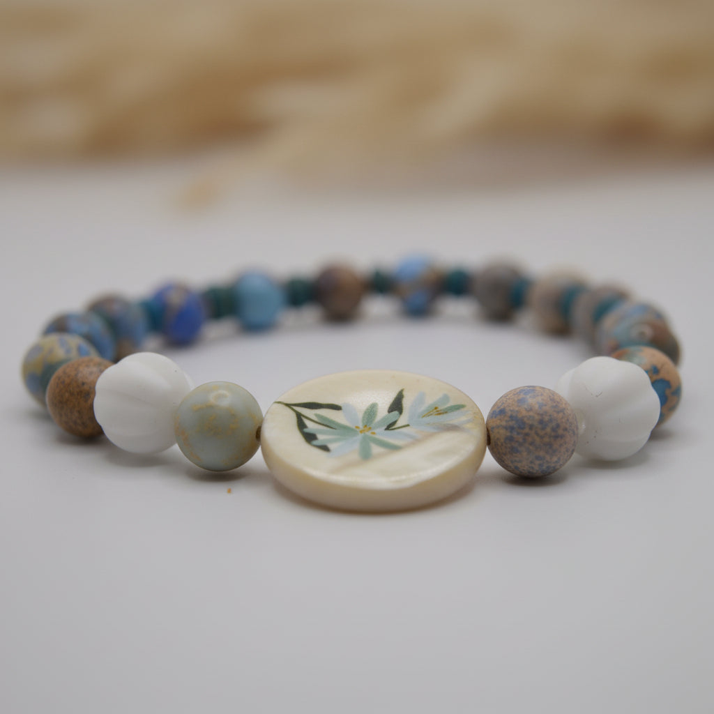 A beaded stretch bracelet featuring a variety of blue, brown, and white beads, with a central round white shell charm decorated with a green bamboo leaf design, set against a plain light gray background.