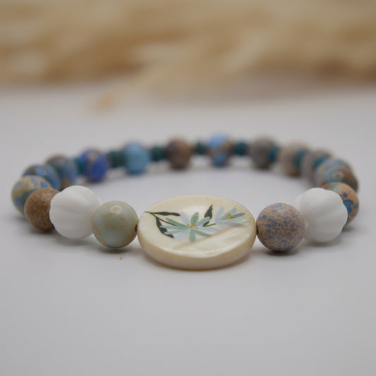 A beaded stretch bracelet featuring a variety of blue, brown, and white beads, with a central round white shell charm decorated with a green bamboo leaf design, set against a plain light gray background.