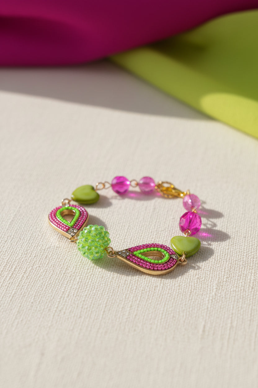 A handmade beaded bracelet with green heart beads, pink rhinestone-encrusted paisley links, and faceted pink and green beads on a gold-tone chain, displayed on a plain light gray surface.