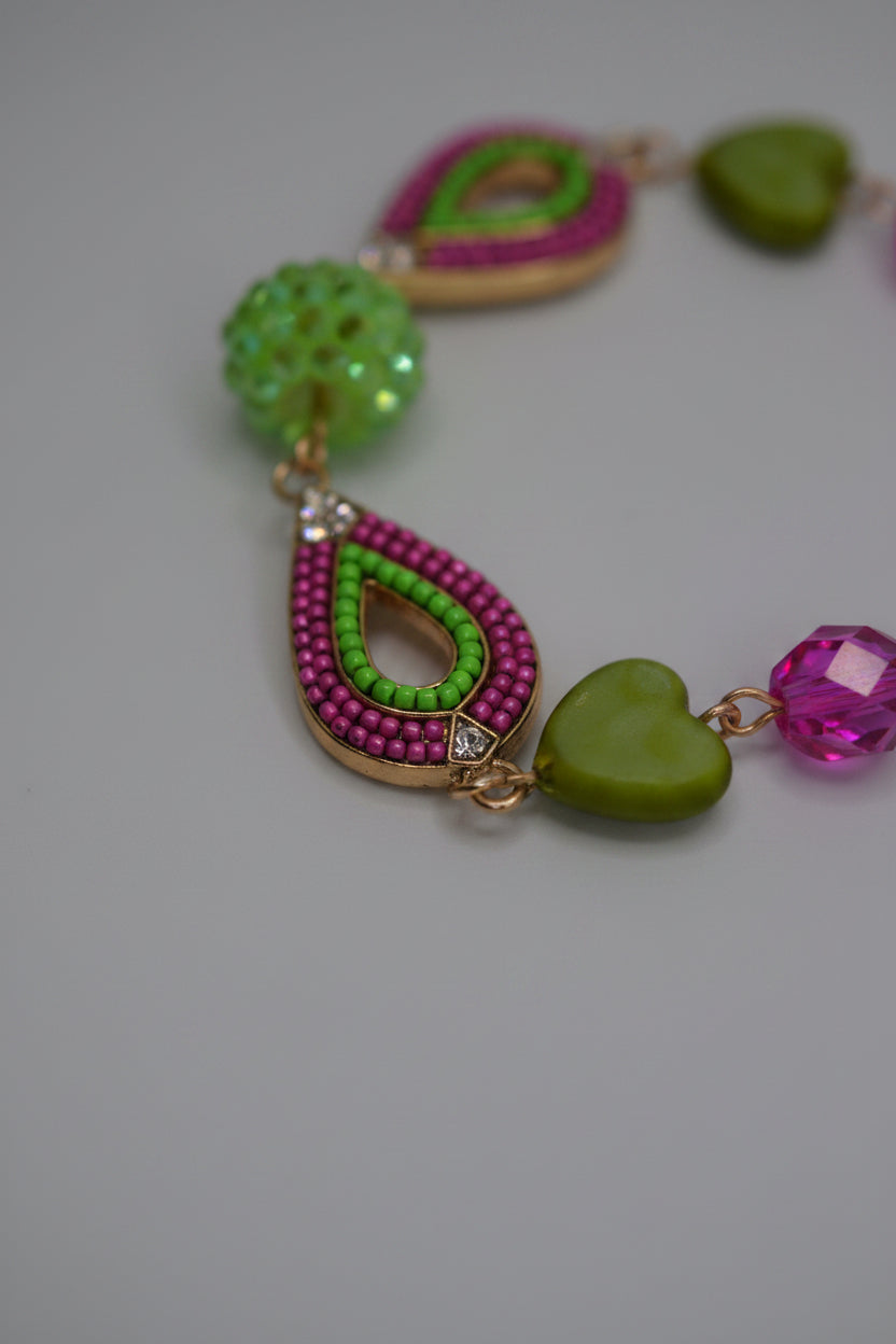 A colorful beaded bracelet with green heart-shaped beads, pink teardrop beads, and gold-toned metal links, displayed on a plain light gray surface.