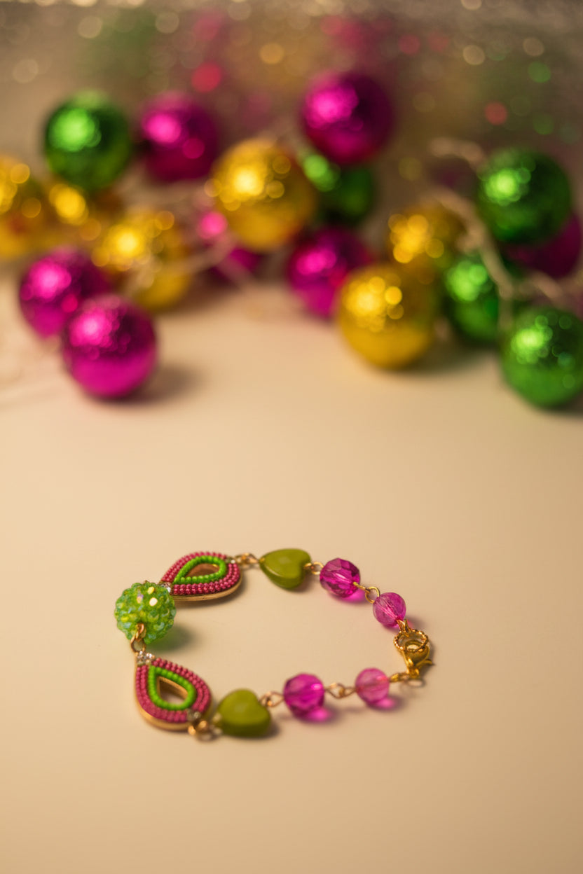 A handmade beaded bracelet featuring green heart-shaped beads, pink flower beads, and pink teardrop beads with gold-toned metal links and a clasp, displayed on a plain light gray surface.