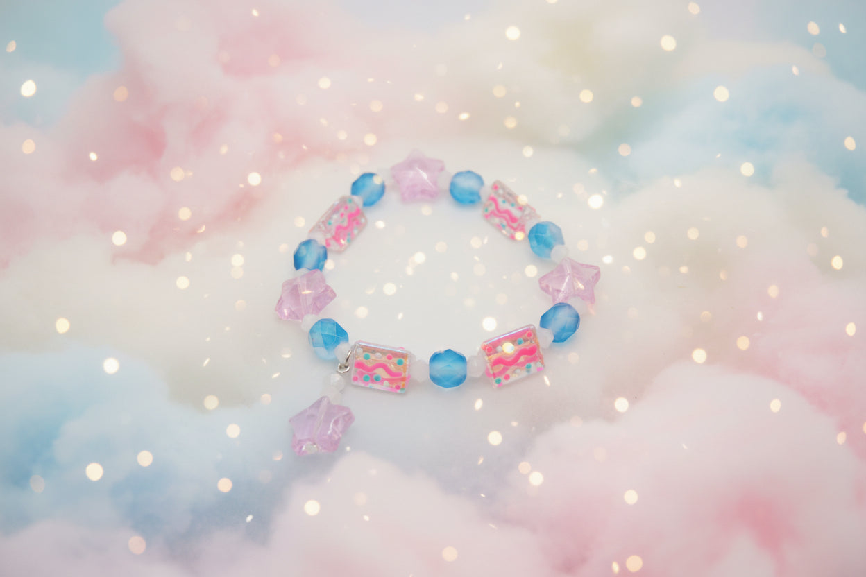 A beaded stretch bracelet featuring pink star-shaped beads, blue faceted beads, and clear rectangular beads with colorful glittery patterns, displayed on a neutral light gray background.