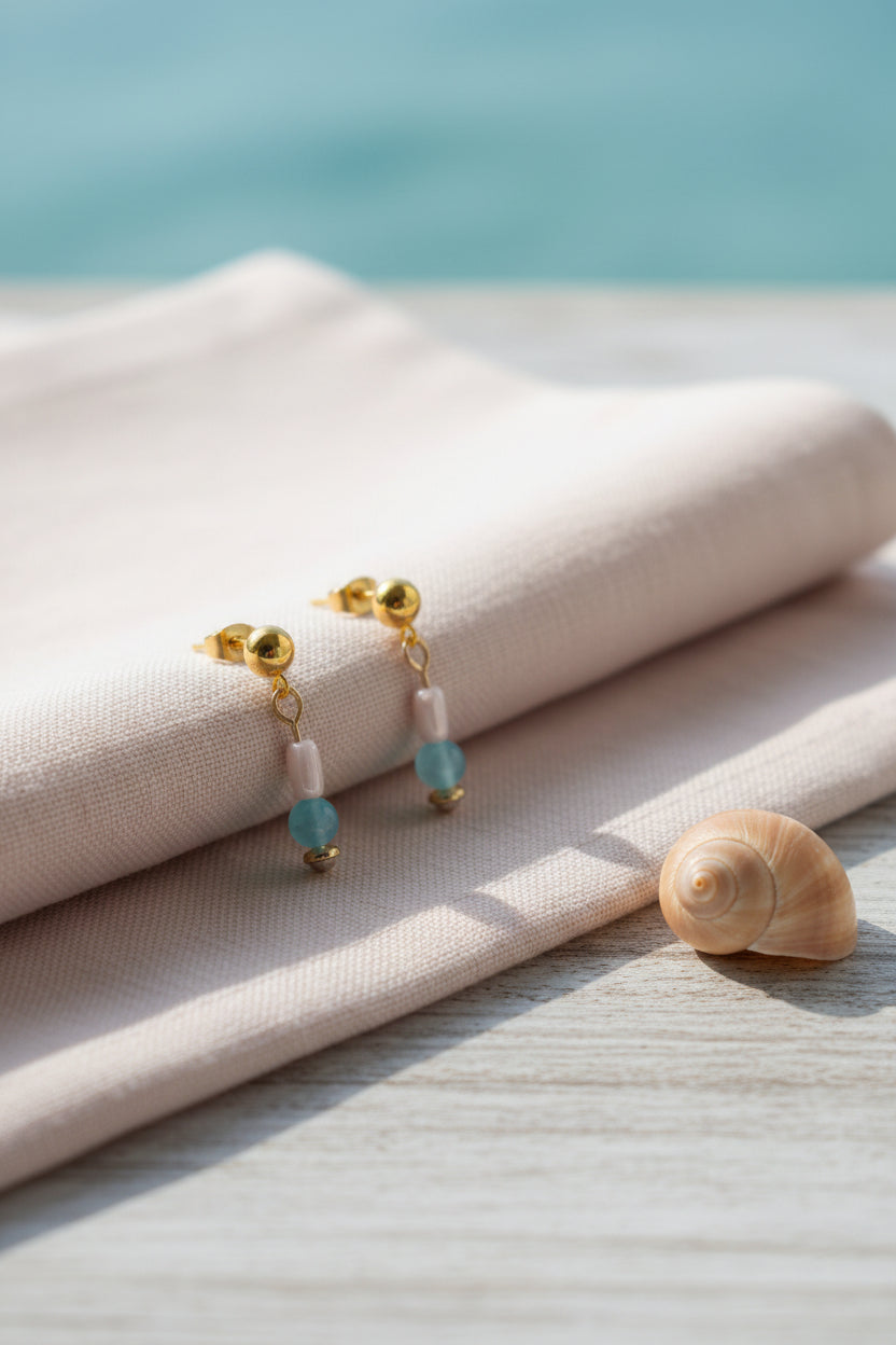 A pair of gold dangle earrings with a light pink cylindrical bead, a light blue round bead, and a small gold cap, displayed on a clear stand against a gray background.