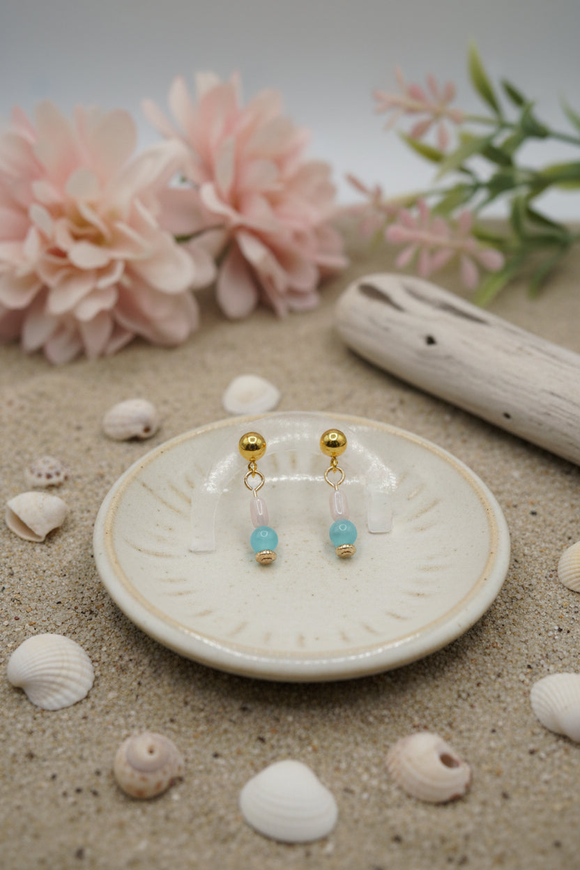 A pair of gold dangle earrings with a light pink bead, a light blue bead, and a small gold disc at the bottom, displayed on a clear stand against a light gray background.