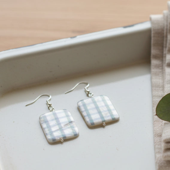 A pair of rectangular drop earrings with a green and beige plaid pattern, suspended from silver fishhook wires, displayed on a clear plastic stand against a neutral gray background.