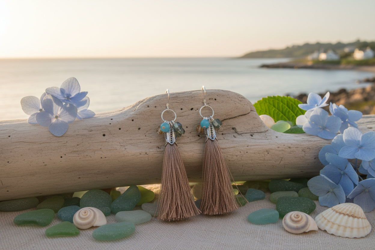 A pair of dangling earrings with silver-toned metal tops, small turquoise blue beads, and long light brown tassel charms, displayed on a clear stand against a light gray background.