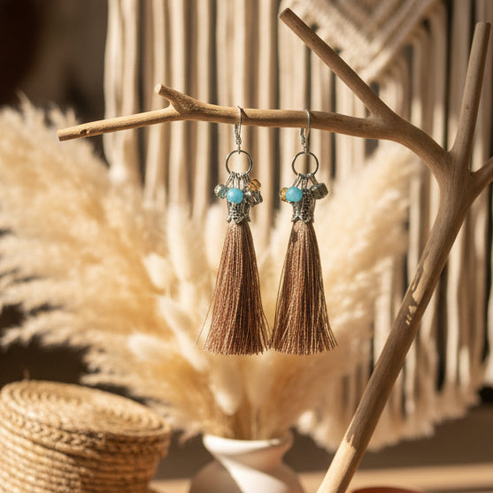 A pair of bohemian-style dangle earrings with silver-toned metal hoops, small turquoise and gold beads, and long brown tassel charms, displayed on a clear stand against a light gray background.