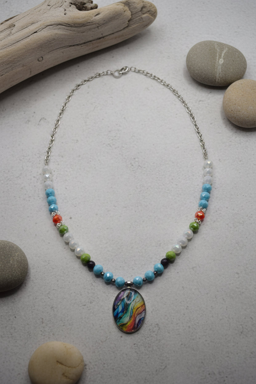A beaded necklace with a colorful oval pendant, displayed on a plain white surface. The necklace features a silver chain, white and clear beads, and small blue, green, orange, and black beads leading to a vibrant, multicolored swirl pendant.